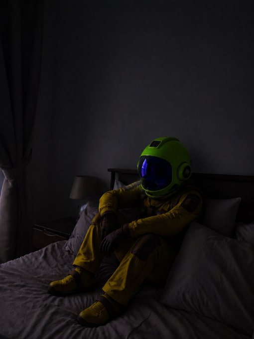 A dimly lit photograph of a person wearing a yellow spacesuit and a green astronaut helmet with a glowing blue visor, sitting on a bed in a dark bedroom.
