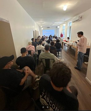 A group of people seated in a room watching a presentation on a projector screen, with a man standing at the front.