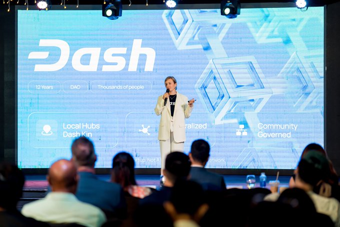 The image shows a woman standing at a podium, giving a speech or presentation to an audience of people who are watching her intently from behind a screen displaying the Dash logo. The event appears to be taking place on a stage with a large screen in front of her, and there is a banner that reads "Onchain Financial Infra" visible in the image as well. The woman's attire consists of a white blazer and pants, suggesting she might be dressed for a formal or professional setting.