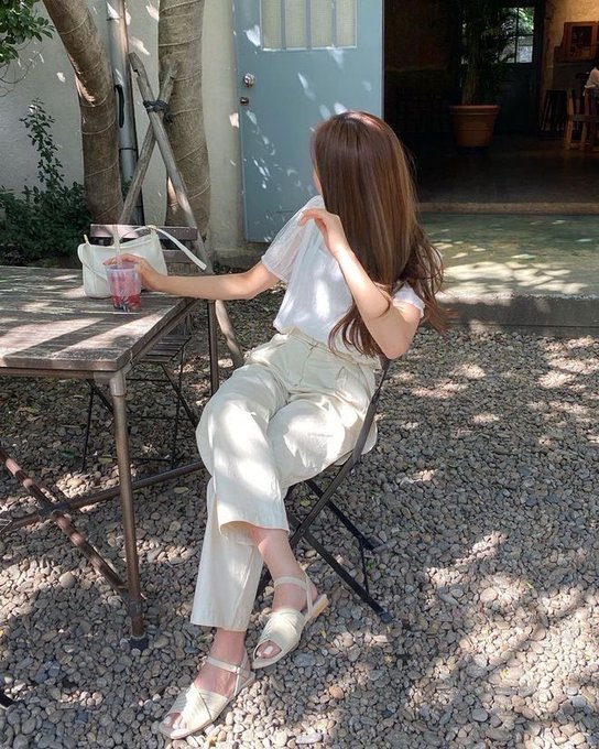 A woman in white clothing sits on a chair at an outdoor cafe table, drinking from a glass in a sunlit courtyard.
