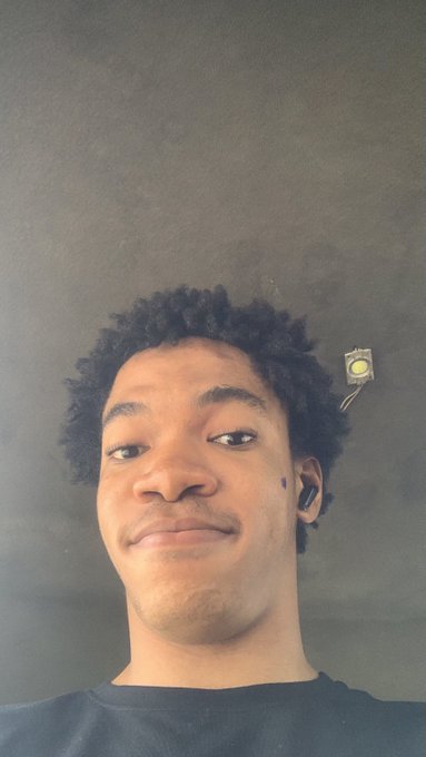 A close-up selfie of a young man with curly hair wearing a black shirt, an earpiece, and what appears to be a small object attached near his ear, against a gray wall background.