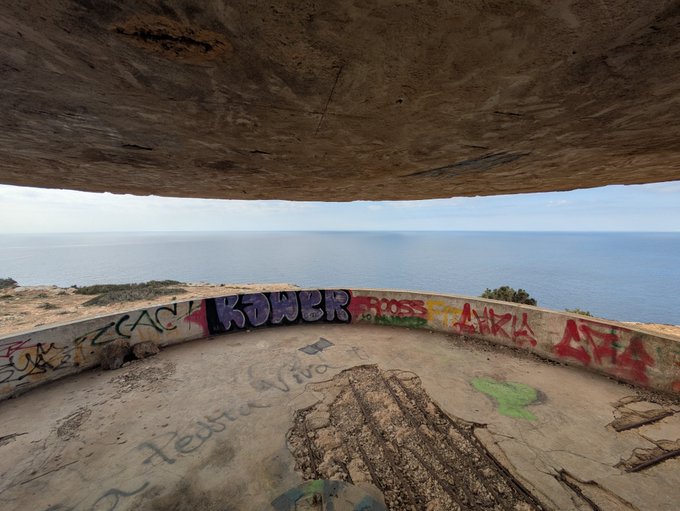 A photograph taken from inside a graffiti-covered concrete bunker or observation post overlooking the sea, with the curved ceiling overhead and names like 'COACH', 'ROSS', 'LARA', and 'Vivo' spray-painted on the walls.
