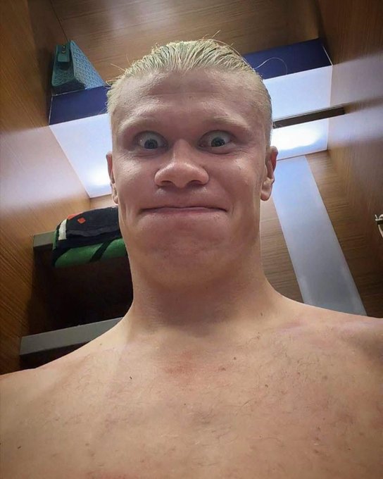 A shirtless selfie of a blonde-haired man making an exaggerated wide-eyed expression in what appears to be a locker room.