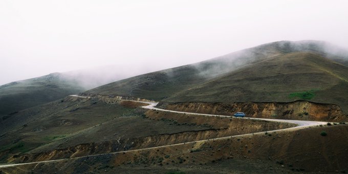 A scenic aerial view of a winding mountain road on a grassy hillside enveloped in fog, with a blue car driving along the curved path.