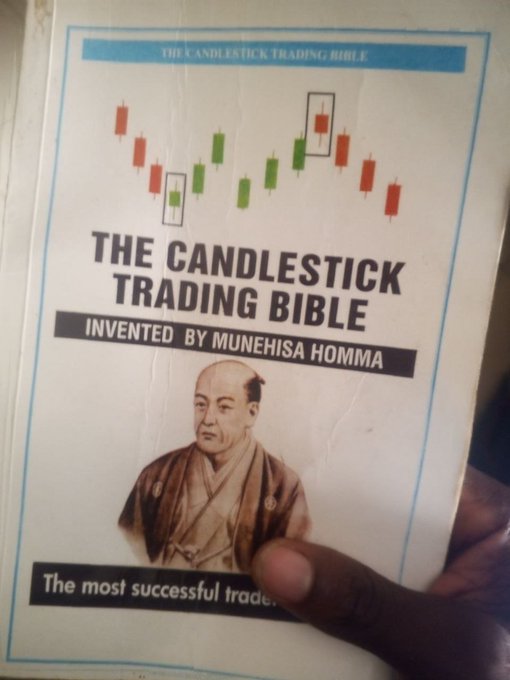 A person holds up a book titled 'The Candlestick Trading Bible: Invented by Munehisa Homma', featuring a portrait of a historical Japanese figure and candlestick chart patterns on the cover.