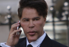 A short video clip thumbnail showing a man in a dark suit and tie holding a silver smartphone to his ear, looking forward with a serious expression outdoors.