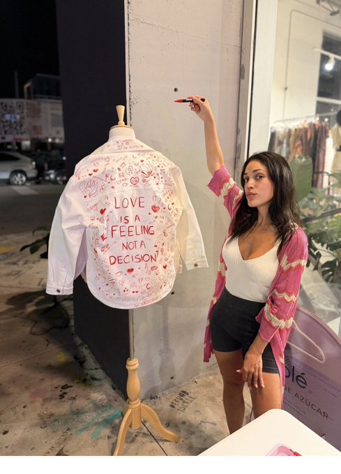 A woman stands next to a white shirt-covered mannequin that reads 'LOVE IS A FEELING NOT A DECISION', holding a marker to add doodles to the shirt.
