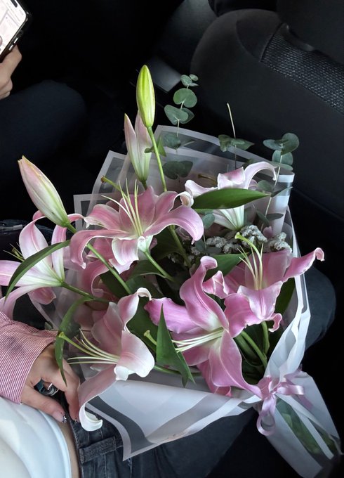 A person holds a bouquet of pink lilies and eucalyptus wrapped in white and black paper inside a car.