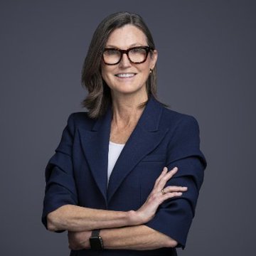 A confident woman with shoulder-length brown hair and black-rimmed eyeglasses smiles with her arms crossed, wearing a navy blue blazer over a white top against a dark gray background. The portrait relates to the post as it depicts Cathie Wood, the founder of ARK Invest, whose firm made the notable $14.2 million Bitcoin purchase.