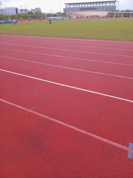A view of a red rubber running track with white lane markers at a stadium, with a green field and buildings in the background.