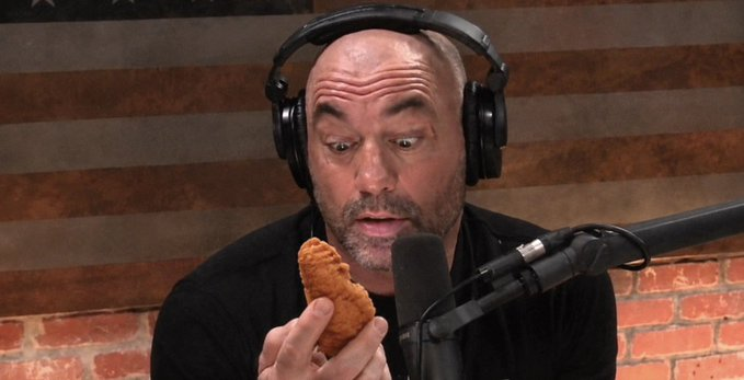 Joe Rogan wearing headphones, holding a piece of fried chicken near a microphone with an American flag in the background.