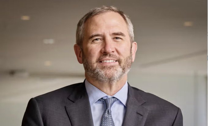 A professional portrait photograph of a middle-aged man with a beard, wearing a dark suit, light blue shirt, and patterned tie.