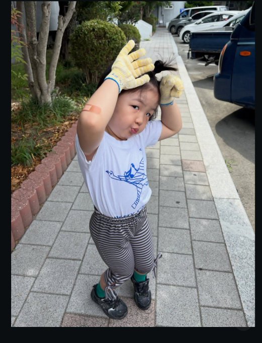 A young child wearing a white t-shirt with a blue design, striped pants, and yellow gardening gloves, posing playfully on a paved sidewalk with cars in the background.