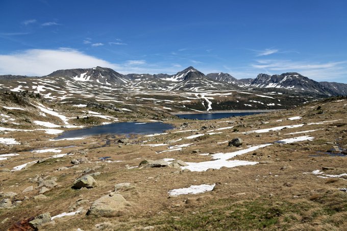 A wide landscape view of a mountainous, snowy environment with scattered rocks, small lakes, and patches of snow under a blue sky with scattered clouds.