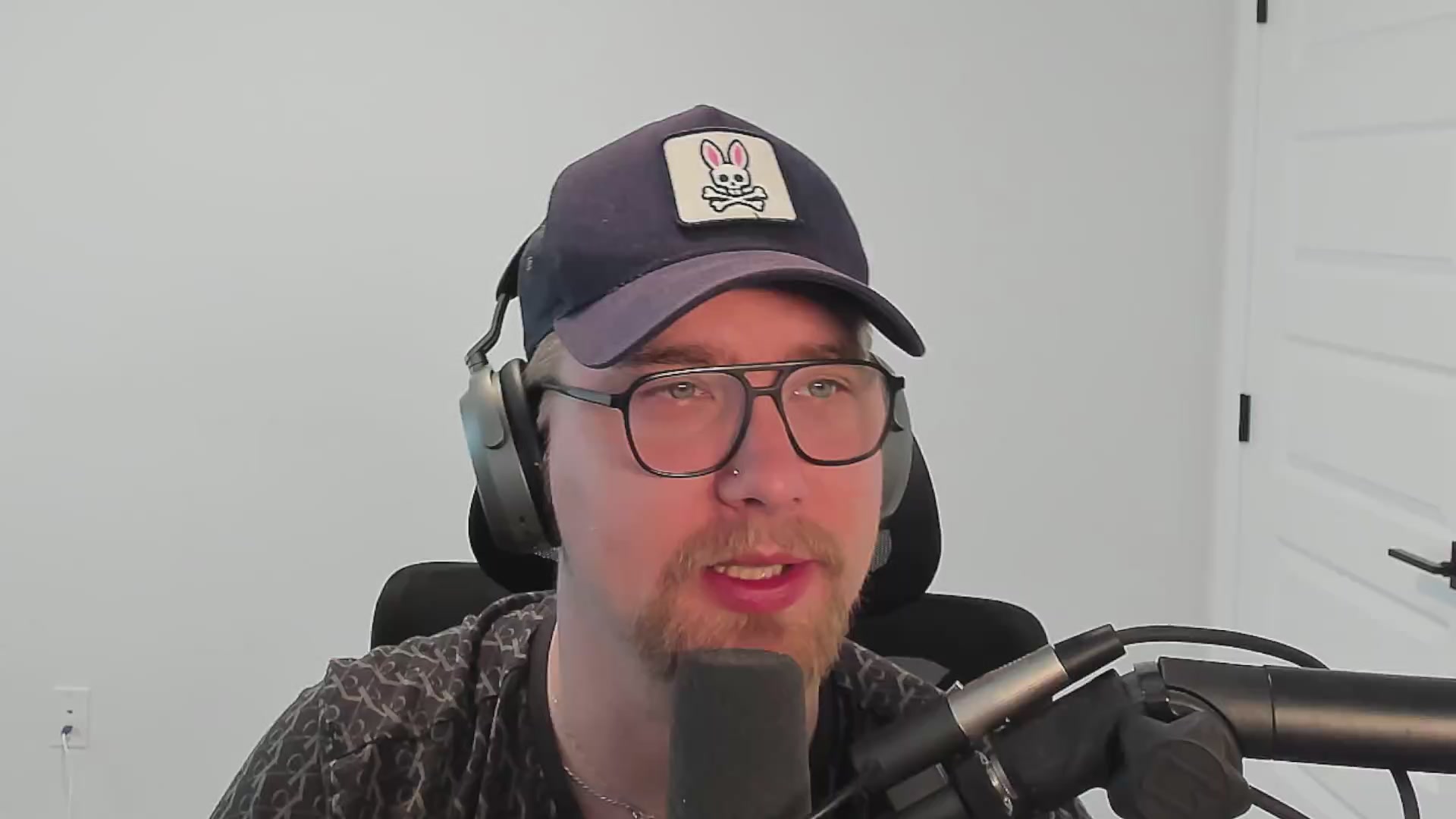 The image features a man wearing glasses, sitting behind a microphone with headphones on his head. He has a hat on that says "bunny ears". The background of the image shows a white wall, which contrasts with the gray floor beneath him. There is also an electrical outlet visible in the scene.
