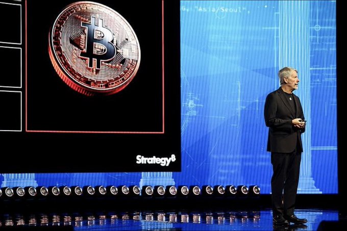 A man in a black suit standing on stage in front of a large Bitcoin logo and 'Strategy' branding, presenting at what appears to be a cryptocurrency or financial conference.