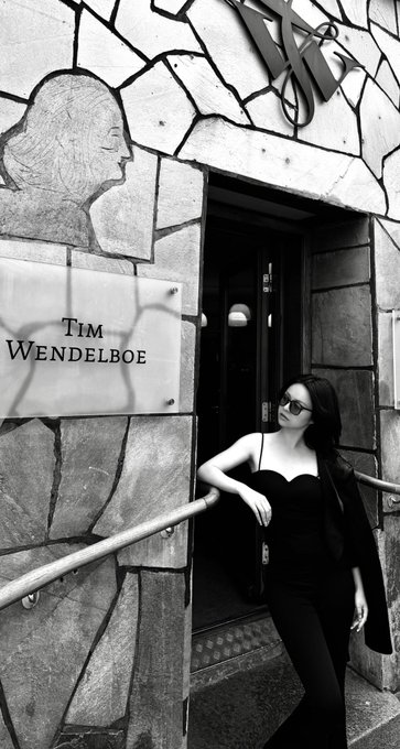 A black-and-white photo of a woman in a black dress and sunglasses standing at the entrance of a building marked with a sign reading 'Tim Wendelboe'.