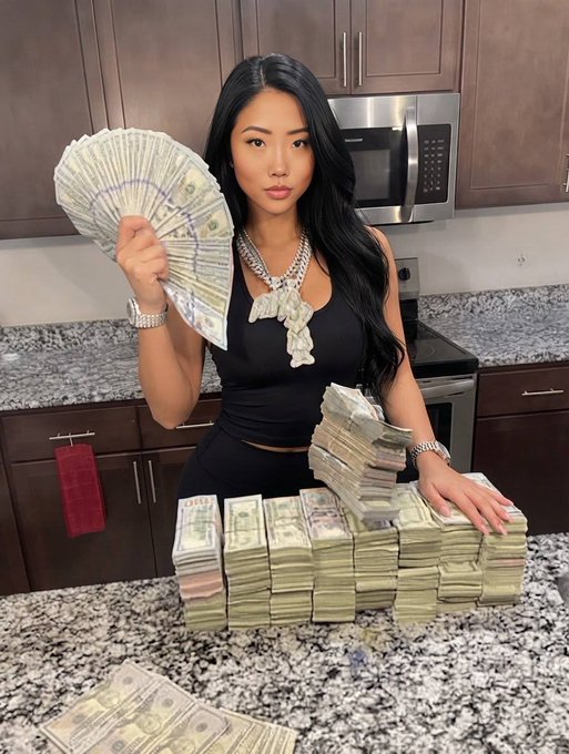 A woman poses in a kitchen surrounded by large stacks of cash, fanning bills in one hand while wearing a chunky diamond chain spelling out a name across her chest. The opulent display of wealth and luxury jewelry fits the caption's tongue-in-cheek jab at how founders flash their success after raising $20M in VC funding.