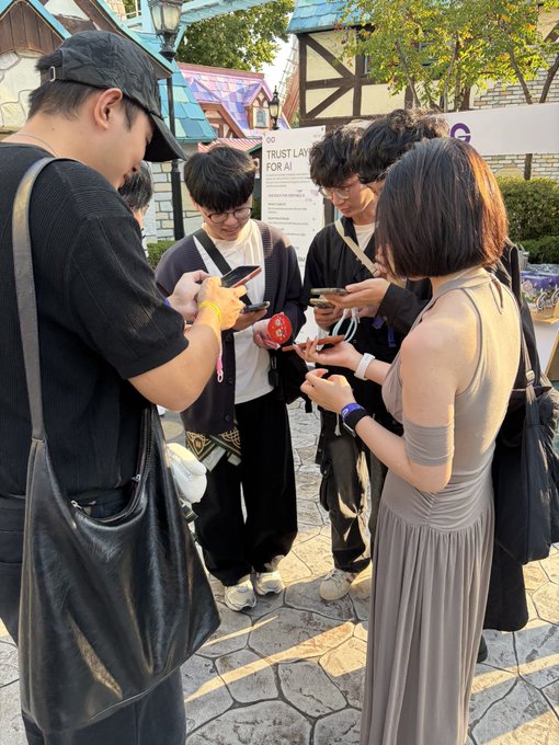 A group of people gathered outdoors at what appears to be an event, looking at their smartphones; one person holds a red 'GMT' token.