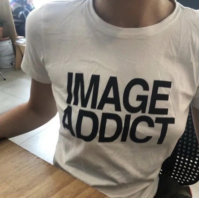 A person wearing an off-white t-shirt with bold black text reading "IMAGE ADDICT" across the chest, photographed indoors near a wooden table.