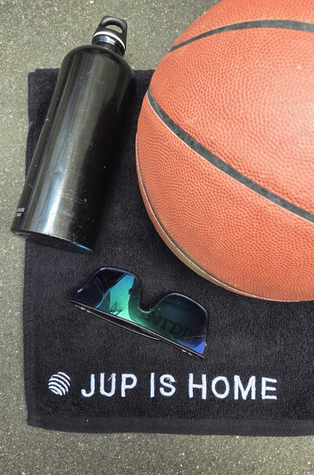 A black water bottle, orange basketball, and teal sunglasses rest on a black 'JUP IS HOME' towel.