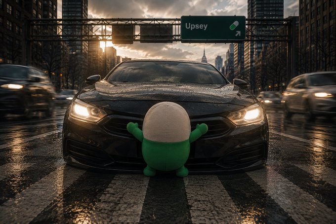 A green and white pill-shaped cartoon character standing in front of a black sports car on a wet city street. A highway sign in the background reads 'Upsey'.
