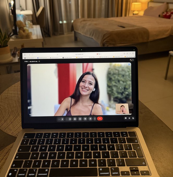 A MacBook laptop displaying a video call with a smiling woman, placed on a wooden table in a modern apartment setting.