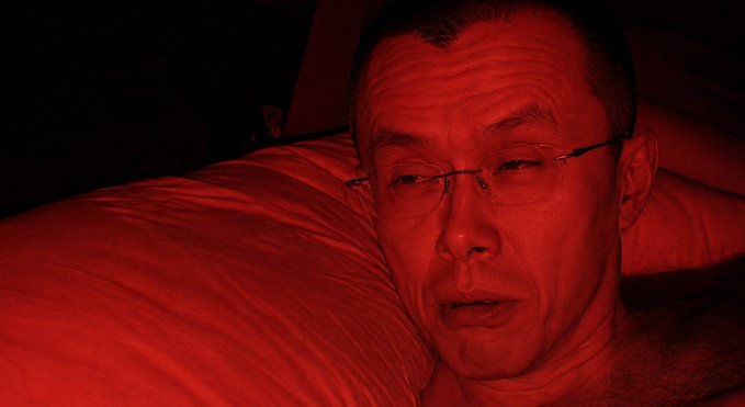 A close-up photo of an Asian man wearing rimless glasses lying down on a white pillow in a dark room illuminated entirely by red lighting.