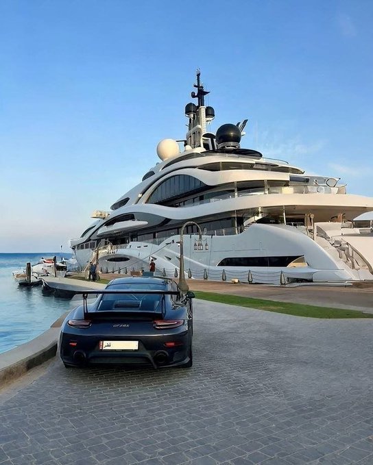 A sleek Porsche GT2 RS parked on a stone-paved dock in front of a massive luxury superyacht moored at a marina under a clear blue sky.