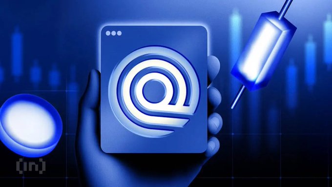 A blue-tinted digital illustration of a hand holding a device with an '@' symbol, with another glowing device attached by a cord against a background of stock market candlesticks.