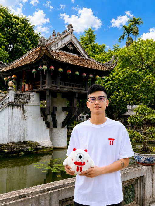 A young man wearing glasses and a white t-shirt holding a plush toy, posing in front of the iconic One Pillar Pagoda, a traditional Vietnamese Buddhist temple in Hanoi surrounded by lush greenery.