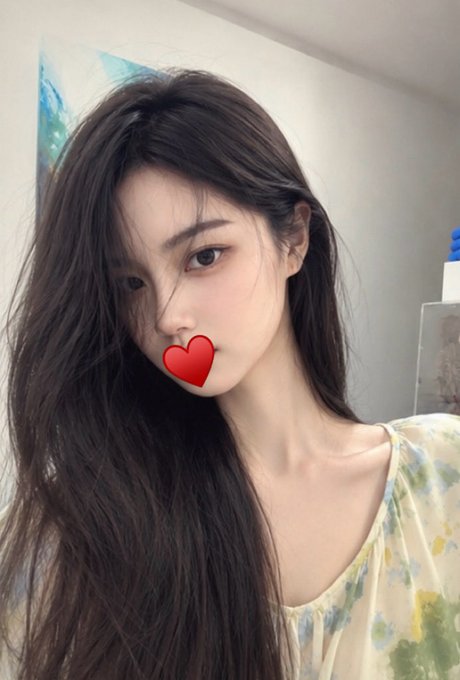 A selfie of a young woman with long dark hair wearing a floral top, with a red heart emoji covering her mouth.