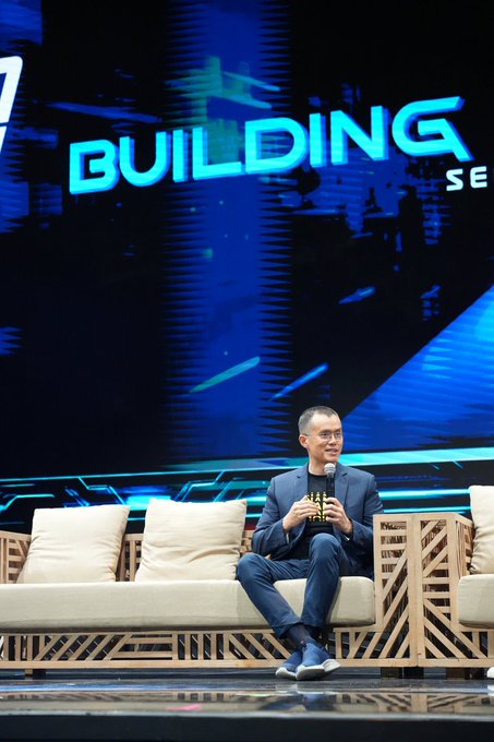 Changpeng Zhao (CZ) sits on a stage couch holding a microphone, with a large screen behind him displaying "BUILDING SE" in bold blue letters. The image captures his first public appearance in the Philippines following his release from prison, as he participates in a panel discussion.
