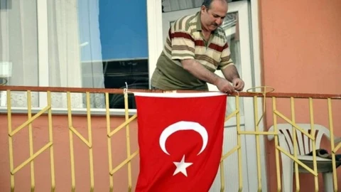 A man stands on a balcony hanging a red Turkish flag with the white crescent and star emblem over the yellow railing.