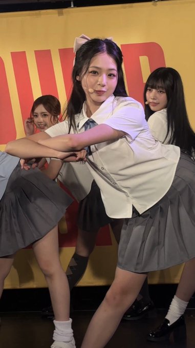 Three young women in matching school-uniform outfits of white tops and gray pleated skirts pose on stage, with the central figure featuring a white bow in her hair.
