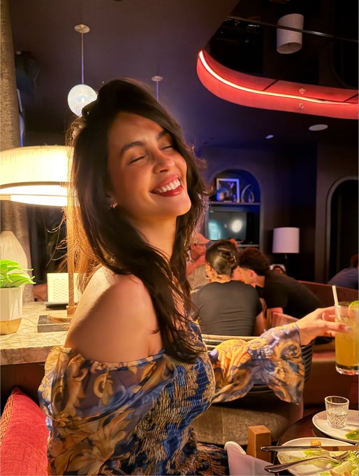 A smiling woman with long dark hair wearing a blue and gold patterned off-shoulder top, holding a glass of orange juice in a dimly lit restaurant with red neon lighting.