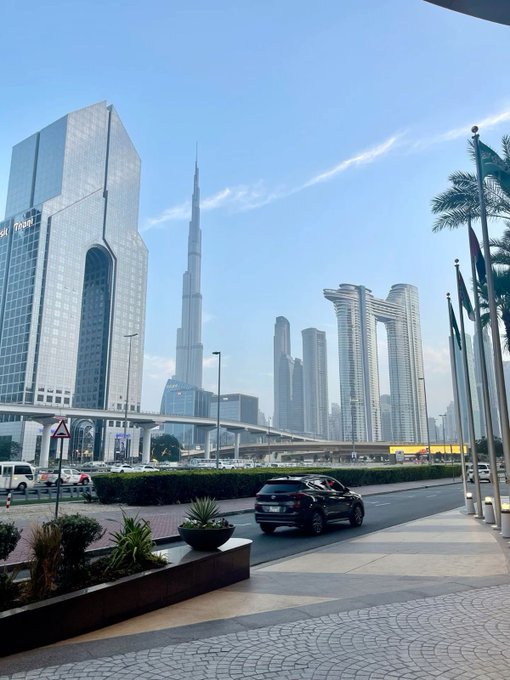 A street-level view of Dubai features the Burj Khalifa and surrounding skyscrapers in the background, with a road, a black SUV, and UAE flags in the foreground.