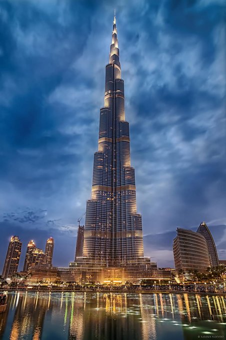 A nighttime photograph of the Burj Khalifa in Dubai, illuminated and reflecting in a pool of water against a dramatic cloudy sky, credited to Karen Kuzniar.