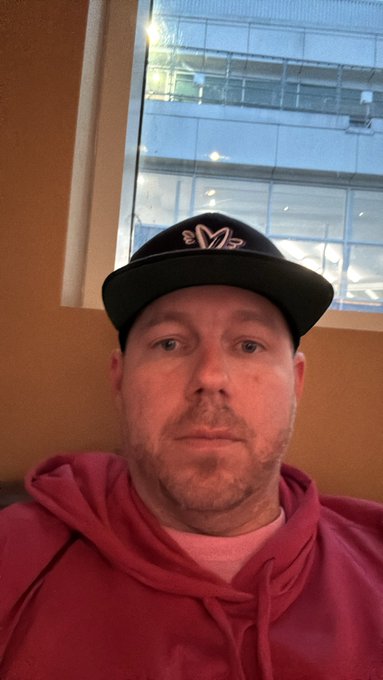 A selfie of a man wearing a pink hoodie and a black baseball cap with a white 'M' logo, sitting indoors with windows visible behind him.