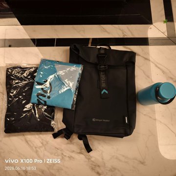 The image features a large package, possibly containing merchandise from a company named "Binance", placed at the entrance of a building. The package appears to be quite large compared to other packages seen in the photo. It's positioned next to a black backpack on a marble floor and a blue water bottle nearby.