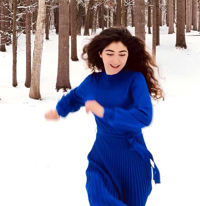 A woman with long curly dark hair wearing a vibrant blue sweater and matching pleated skirt dances joyfully in a snowy forest.