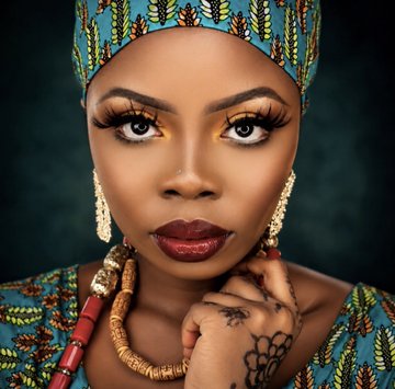 The image features a woman with dark skin wearing a turquoise headband, red earrings, and a gold necklace. She has her hand resting on her chin while looking directly at the camera. The background of the photo is black, which contrasts with the colorful elements in the foreground. The caption below the image reads "Amina represents stability within Ancestra. Through Amina you’re connecting RITUAL to stable assets designed for consistency and smoother trading experiences Amina simply offers the calmest route forward to swap Ritual to stables Stable Mode GM GM ancestra-dex.vercel.app".