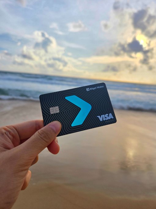 A hand proudly displays a sleek black "Bitget Wallet Visa" card with a bold blue arrow design, set against a serene beach sunset. The image symbolizes the upward trajectory and global reach of Bitget's expansion, reinforcing the post's message of unstoppable growth and widespread adoption.