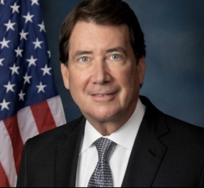 An official portrait of a man in a dark suit and patterned tie, positioned in front of an American flag against a blue background.