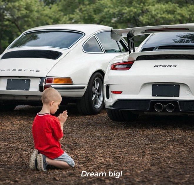 A young boy in a red shirt kneels on the ground with hands clasped in prayer, gazing at two white Porsche cars (a classic 911 and a modern GT3RS) with the caption 'Dream big!' at the bottom.