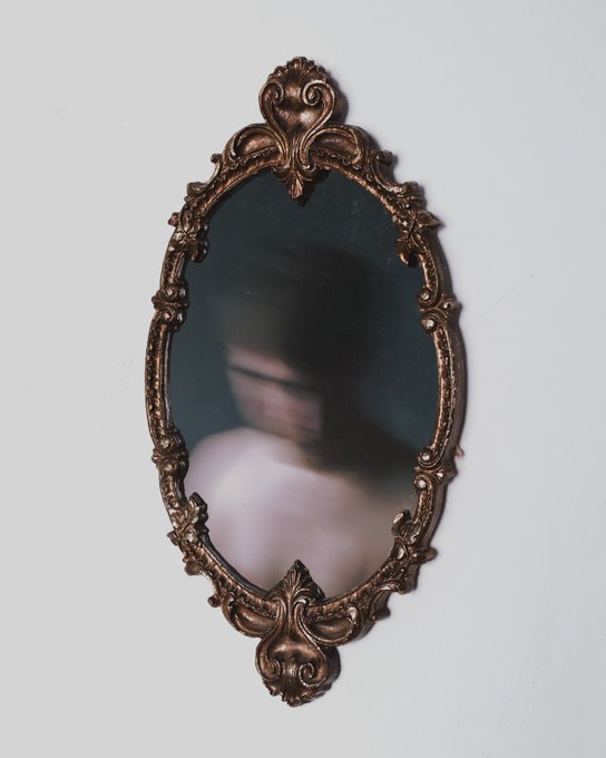An oval, ornately carved copper/bronze vintage mirror frame mounted on a plain white wall, with a blurry reflection visible in the glass.