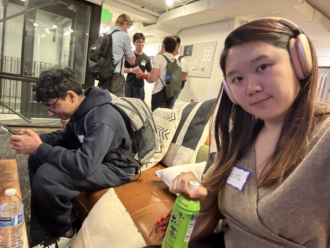 A casual photo of a young woman wearing headphones sitting on a couch holding a green drink can, with a name sticker visible, and people in the background.