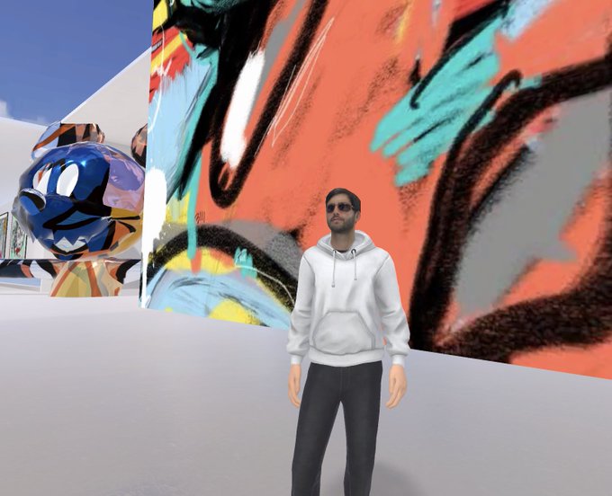 A digital avatar of a man wearing sunglasses, a white hoodie, and black pants stands in a virtual environment in front of a large, colorful abstract mural.