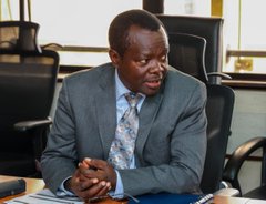 A man in a dark suit and patterned tie sits at a desk in an office, appearing to be in a meeting or interview.