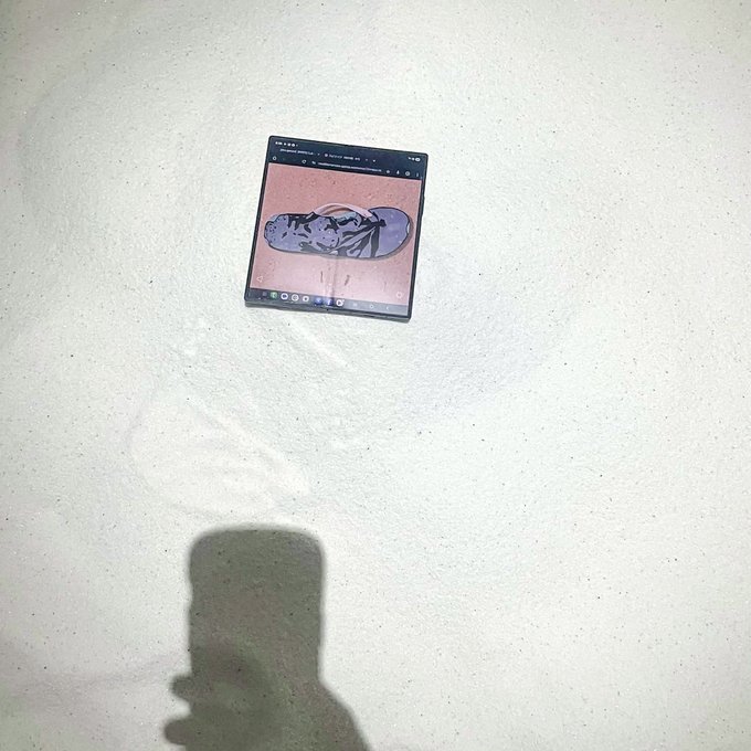 A foldable smartphone lies on a sandy surface, displaying an image of a skateboard on its screen, with the photographer's shadow visible in the foreground.